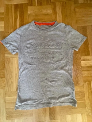 Grå t-shirt från Superdry - Säljer en grå t-shirt från Superdry med broderad logotyp på bröstet och diskret text. T-shirten har korta ärmar och rund halsringning med orange detalj på insidan. Perfekt för dig som gillar stilrena plagg med subtil branding.