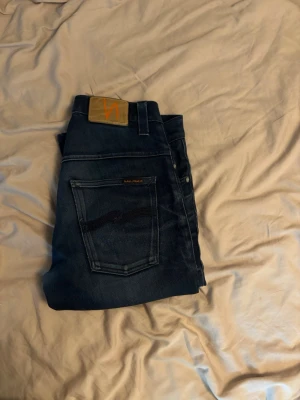 Mörkblå jeans från Nudie Jeans - Snygga mörkblå jeans från Nudie Jeans med klassisk femficksdesign och subtila slitningar. Jeansen har raka ben och är tillverkade i denim med en skön känsla. Perfekta för en avslappnad och trendig stil.