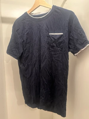 Mörkblå t-shirt från Erla of Sweden - Snygg mörkblå t-shirt från Erla of Sweden med vit detalj vid halsringningen och en liten vit kant på bröstfickan. Klassisk passform och korta ärmar, perfekt till jeans eller shorts. Tillverkad i mjuk bomull för skön känsla.