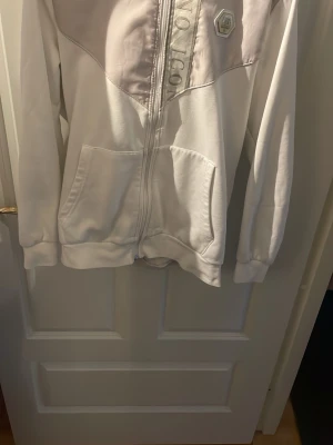 Vit hoodie från icons lyxary - Snygg vit hoodie från Juicy Couture med dragkedja framtill och stora fickor. Har en huva och ribbade muddar. Märkets logga syns på bröstet och längs nederkanten står det 'Juicy Couture'. Perfekt för en chill och trendig look.