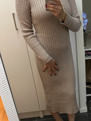 Beige ribbad stickad långklänning - Snygg beige ribbad stickad långklänning med lång ärm och hög hals. Klänningen har en kroppsnära passform och är perfekt för dig som gillar en stilren och enkel look. Supermjuk och skön att bära.
