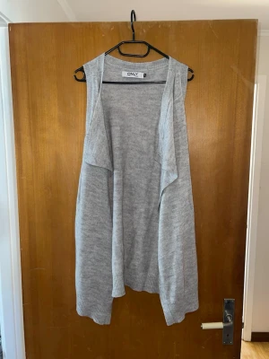 Ljusgrå stickad väst från ONLY - Säljer en ljusgrå stickad väst från ONLY. Västen är ärmlös, har öppen front och mjukt fall. Perfekt att slänga över en långärmad tröja för en chill och snygg look. Materialet är mjukt och skönt, och ribbstickade kanter ger extra stil.