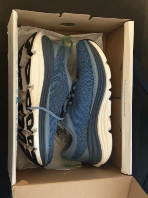 Blå Hoka sneakers med tjock sula - Snygga blå sneakers från Hoka med mönstrad ovandel i mesh och tjock, vit sula med svart detalj undertill. Skorna har gröna detaljer på hälen och klassisk snörning. Perfekta för dig som gillar sportig stil och komfort.