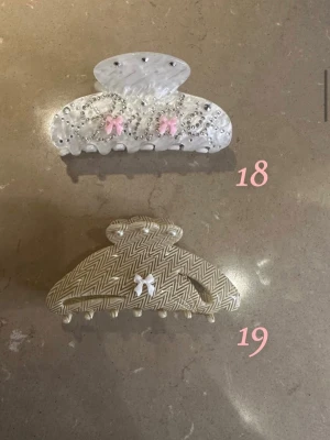 Två hårklämmor med rosetter, pärlor, diamanter och mönster - Säljer två hårklämmor, 1 för 89 2 för 165. Den ena är vit med silverglittrande och pärlor detaljer och rosa rosetter, den andra har ett beige och vitt zigzag-mönster med en liten vit rosett. 