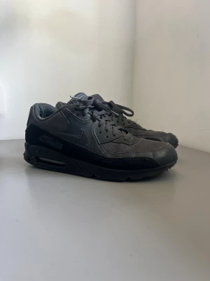 Nike Air Max 90 svarta sneakers - Nike Air Max 90 sneakers i helsvart med diskret Swoosh-logga på sidan och Air Max-detalj vid sulan. Ovandelen är i en mix av läder och textil, med klassisk synlig luftbubbla i sulan. Snygg och tidlös modell med robust känsla och snörning framtill.