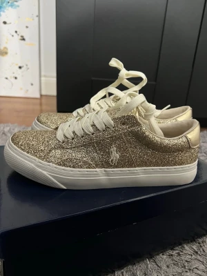 Glittriga guldiga sneakers från Polo - Snygga sneakers från Polo Ralph Lauren i glittrigt guld med vita snören och vit sula. Låg modell med klassisk Polo-logga på sidan. Perfekta för dig som vill sticka ut med en trendig och glamorös look. Materialet är syntet med glitterfinish. Skorna är helt nya och aldrig använda, har kvar boxen och kvittot, skorna är i storlek 35 men passar ändå mig som har storlek 36/37