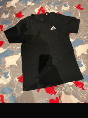 Svart Adidas t-shirt med logga - Svart t-shirt från Adidas med klassisk vit logga på bröstet. Enkel och stilren design med rund hals och korta ärmar. Perfekt för en sportig och avslappnad look.