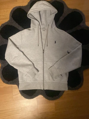 Grå zip hoodie från Polo Ralph Lauren - Snygg ljusgrå hoodie med dragkedja från Polo Ralph Lauren. Klassisk design med huva och dragsnören, ribbade muddar och fickor framtill. Liten broderad logga på bröstet. Perfekt till chill vardagsstil. Mjuk bomullsblandning för extra komfort.
