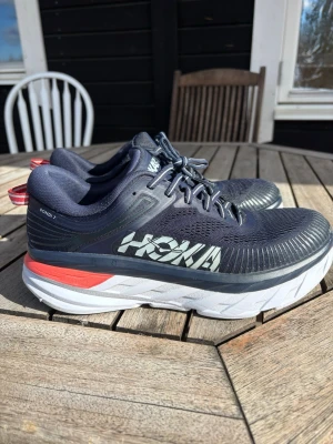 Hoka Bondi 7 mörkblå sneakers - Snygga Hoka Bondi 7 sneakers i mörkblå mesh med vit och röd detalj på sulan. Skorna har en chunky, vadderad sula för extra komfort och stora Hoka-loggor på sidan. Perfekta för dig som gillar streetwear och sportig stil.
