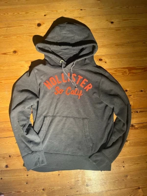 Vintage Hollister hoodie   - Sällsynt och snygg Hollister hoodie i mörkgrå färg med orange text. Tröjan är i storlek L men passar snarare S-M. Tröjan är i mycket bra skick utan några som helst defekter, skriv vid eventuella funderingar😄