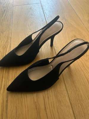 Svarta slingback pumps med smal klack - Stilrena svarta pumps med spetsig tå och smal klack. Skorna har öppen häl med slingback-rem och är tillverkade i mockaimitation. Ca 7-8 cm klack. Andvända en gång.