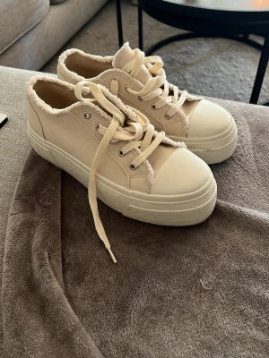 Beige sneakers från Even&Odd - Stilrena beige sneakers från Even&Odd med platåsula och råa kanter. Skorna har en canvasliknande ovandel, vita snören och tåhätta i gummi. Perfekta för dig som gillar en clean och trendig look med lite extra höjd.