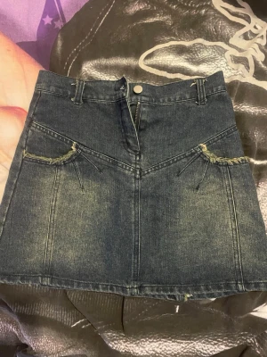 Mörkblå jeanskjol med fransdetaljer - Säljer en snygg mörkblå jeanskjol med fransade detaljer längs fickor och sömmar. Kjolen har en kort modell, bälteshällor och knappstängning framtill. Säljer pågrund av att jag inte har användning för den, skriv vid frågor eller så!