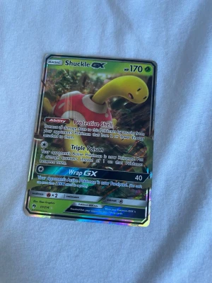 Shuckle GX Pokémonkort holografiskt - Shuckle GX holografiskt Pokémonkort med färger i gult, rött och grönt. Kortet har glansig yta och visar Shuckle i en naturmiljö. Text och effekter på engelska. Perfekt för samlare eller Pokémonfans.