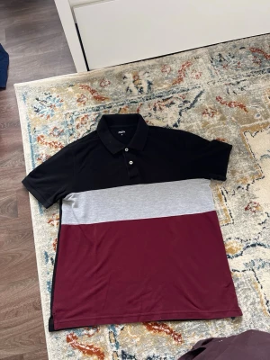 Färgblockad tröja  - Snygg tröja  med färgblock i svart, grått och vinrött. Tröjan har klassisk krage, korta ärmar och knappar framtill. Perfekt för en stilren och avslappnad look.