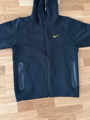 Svart  NOCTA hoodie med dragkedja - Svart hoodie från  NOCTA med hel dragkedja, två vattentäta fickor med dragkedja och huva. Gult Nike Swoosh på bröstet och NOCTA-tryck på ärmen. Sportig och clean design, perfekt för streetwear. Materialet är mjukt och stretchigt. Liten i storlek 