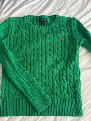 Grön kabelstickad tröja Ralph Lauren - Snygg grön kabelstickad tröja från Ralph Lauren med rund halsringning och lång ärm. Tröjan har det klassiska rosa broderade Polo-loggan på bröstet och breda ribbade muddar vid ärmslut och nederkant. Perfekt för dig som gillar preppy stil.
