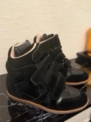 Svarta high top sneakers med kardborre - Snygga svarta high top sneakers med flera kardborreband och gummisula. Skorna har en cool design i mocka och läderimitation, med vit insida och detaljer som ger en sportig vibe. Perfekta för dig som vill sticka ut med din stil.