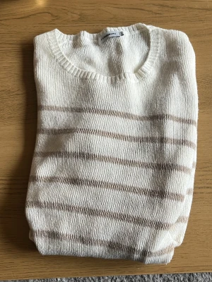 Beige och vit randig stickad tröja - Snygg stickad tröja i beige och vitt med horisontella ränder från J Lindberg . Tröjan har rund halsringning, långa ärmar och ribbade muddar vid ärmslut och nederkant. Perfekt för en avslappnad och trendig look.