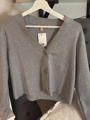 Grå croppad kofta från H&M - Snygg grå croppad kofta från H&M med v-ringning och stora knappar framtill. Koftan har lång ärm och är tillverkad i mjuk bomullsmix, perfekt att slänga över en topp eller klänning för en chill look.