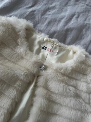 Vit pälsväst från H&M med kristallknapp - Superfin vit pälsväst från H&M i mjuk fuskpäls med horisontella ränder och glittrig kristallknapp framtill. Västen har rund halsringning och är fodrad med slätt tyg. Perfekt för att lyfta din outfit med en lyxig touch.