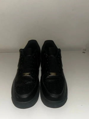 Nike Air Force 1 Low helsvart läder - Säljer ett par klassiska Nike Air Force 1 Low i helsvart läder. Skorna har perforerad tå, svart sula och svarta skosnören med gulddetaljer. Perfekt för dig som gillar en clean och tidlös sneaker med streetkänsla.