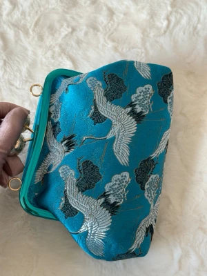 Blå handväska med tranor - Unik blå handväska med broderade tranor och moln i silver och svart. Väskan har metallram i matchande turkos och guldfärgade detaljer. Materialet är glansigt tyg med asiatiskt inspirerat mönster. Perfekt för dig som vill sticka ut med en färgstark accessoar.