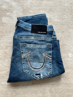 Blå True Religion jeans med broderi - Säljer ett par blå jeans från True Religion med ikonisk broderad bakficka och snygga slitningar. Jeansen har rak passform och klassisk femficksdesign. Perfekta för dig som gillar streetwear och vill ha ett par jeans med attityd.