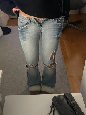 Ljusblå slitna bootcut jeans - Säljer ett par ljusblå bootcut jeans med slitningar och hål på knäna. Jeansen har en klassisk femficksmodell och detaljerade sömmar på bakfickorna. Perfekta för en avslappnad och trendig look. Två skärphållare trasiga