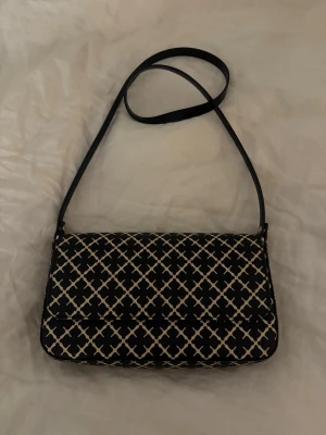 Svart axelväska med vitt mönster - Snygg svart axelväska med vitt rutmönster och lång axelrem från Malene Birger. Väskan har ett lock med magnetknäppning och ett innerfack med dragkedja. Insidan är beige och materialet känns som konstläder. Perfekt för dig som gillar stilrena accessoarer.