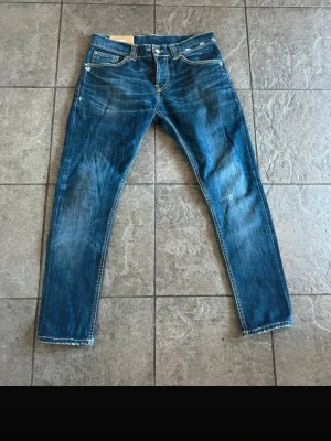 Blå jeans med slitningar dondup - Säljer ett par blå jeans dundup med snygga slitningar och kontrastsömmar. Jeansen har klassisk femficksmodell, dragkedja och knapp i midjan samt detaljerade bakfickor med dragkedjor. Perfekta för en avslappnad och trendig look.