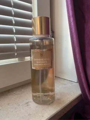 Shimmering Shores body mist 250 ml - Fräsch body mist från Victoria's Secret i doften Shimmering Shores med inslag av coconut sand och driftwood. Flaskan är genomskinlig med guldigt lock och etikett. Innehåller 250 ml och har en elegant, cylinderformad design.