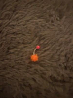 Piercingsmycke med orange boll - Piercingsmycke med böjd stav i metall, dekorerad med en orange fluffig boll och en rosa kula. Perfekt för dig som vill ha något färgglatt och lekfullt till din piercing. Passar till exempel navelpiercing.
