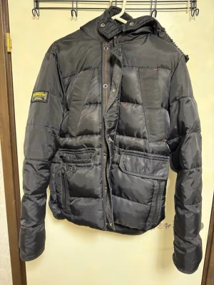Svart pufferjacka Parajumpers - Svart pufferjacka från Superdry med huva, stora framfickor och patch på ärmen. Jackan har dragkedja och knappar framtill samt detaljer i tyg på bröstet. Perfekt för kalla vinterdagar och riktigt snygg streetstil.