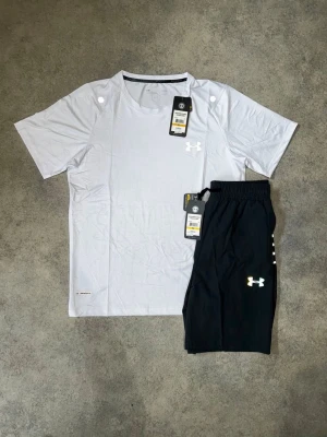 Under Armour träningsset t-shirt & shorts - Snyggt träningsset från Under Armour med en vit t-shirt och svarta shorts. T-shirten har rund hals och kort ärm, medan shortsen har resår i midjan och logga på benet. Perfekt för gymmet eller löprundan.