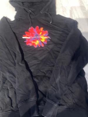 Svart hoodie från Vans med blomtryck - Svart hoodie från Vans med färgglatt blomtryck på bröstet och klassisk huva med dragsko. Mjuk insida och känguruficka framtill. Perfekt för dig som gillar streetwear och vill ha något unikt i garderoben.