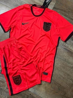 England röd fotbollsdräkt Nike - Säljer ett rött fotbollsset från Nike med England-emblem på både tröja och shorts. Setet har svarta detaljer vid krage, ärmslut och längs shortsen. Tillverkat i lätt och ventilerande material, perfekt för träning eller match.