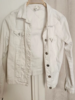 Vit jeansjacka i klassisk modell - Snygg vit jeansjacka med klassisk krage och silverfärgade knappar. Jackan har två bröstfickor med lock och knapp samt raka ärmar. Perfekt att slänga över en t-shirt för en fräsch look. Materialet är jeans och jackan har en tidlös passform.