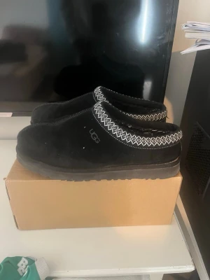 Svarta UGGs Tasman tofflor  - Säljer ett par svarta UGG tofflor i mocka med tjock sula och vit broderad kant runt öppningen. Klassisk slip-on modell som är både stilren och bekväm. Perfekt för att chilla hemma eller snabbt dra på sig när man ska ut med soporna.