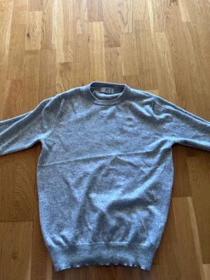 Grå rundhalsad tröja från Bvaldi - Snygg grå tröja från Bvaldi med rund halsringning och diskret broderad logga på bröstet. Tröjan har ribbade muddar vid ärmslut och nederkant, vilket ger en klassisk och stilren look. Perfekt att matcha med jeans eller chinos.