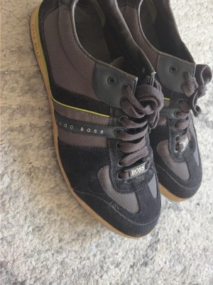 Hugo Boss skor - Lite smutsiga men annars i bra skick, behövs en tvätt/rengöring bara, Snygga svarta sneakers från Hugo Boss med detaljer i mocka och mesh. Skorna har en diskret limegrön rand, svarta snören och gummisula. Märkeslogga på sidan och tungan. Perfekta för en trendig streetwear-look.