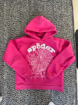 Sp5der hoodie spider hoodie - Cool rosa hoodie med vit spindelnätsprint och texten 'spider' dekorerad med små glittrande stenar. Hoodien har en stor huva och ribbade muddar. Perfekt för dig som gillar unika och färgstarka plagg.