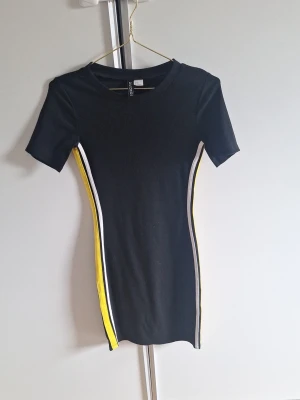 Svart kortklänning från H&M Divided - Säljer en svart kortklänning från H&M Divided med sportiga ränder i vitt och gult längs sidorna. Klänningen har en tight passform och korta ärmar. Nopprar av innan frakt. Katt finns i hemmet