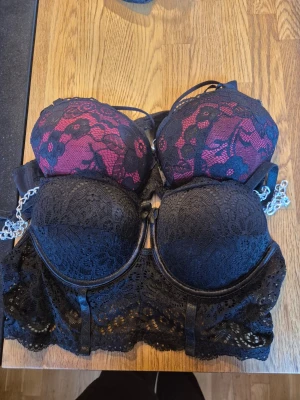 Två spetsbhar i svart och vinrött - Säljer två bhar med bygel och vaddering, båda i svart spets. En har vinröd bas med svart spets ovanpå och den andra är helsvart med längre spetskant nedtill. Båda har justerbara axelband och hakknäppning bak. Ena är med push up och den andra utan.    Alla kläder rollas innan dom skickas hem då det finns katt i hemmet🐈 Jag stryker och tvättar INTE innan jag skickar det får du stå för själv så det blir som du vill ha det🤗