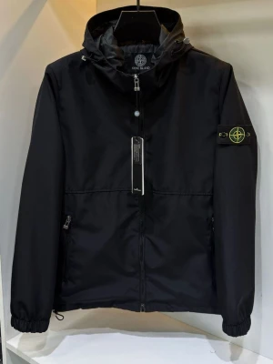 Stone Island Windbreaker - Svart vindjacka från Stone Island med huva och dragkedja framtill. Jackan har två sidofickor med dragkedja och den klassiska logotypen på vänster ärm. Materialet är lätt och vindtätt, perfekt för blåsiga dagar.