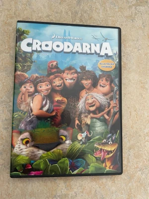 DVD Croodarna DreamWorks svensk tal - DVD-film Croodarna från DreamWorks med svenskt tal. Omslaget har färgglada illustrationer av animerade grottmänniskor och fantasidjur. Innehåller extramaterial som grottdagbok och borttagna scener. Perfekt för dig som gillar animerade familjefilmer.