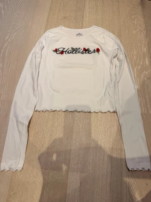 Vit croppad långärmad topp Hollister - Vit ribbad croppad långärmad topp från Hollister med broderad logga och röda rosor framtill. Söt volangkant vid ärmslut och nederkant. Perfekt för dig som gillar en feminin och trendig stil.