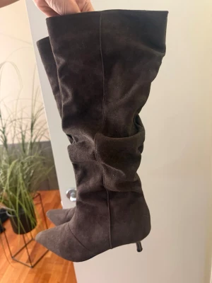 Bruna höga mockastövlar med klack - Snygga bruna stövlar i mocka med hög skaft och smal klack. Stövlarna har en slouchy design och spetsig tå, vilket ger en trendig och elegant look. Perfekta för att lyfta vilken outfit som helst.