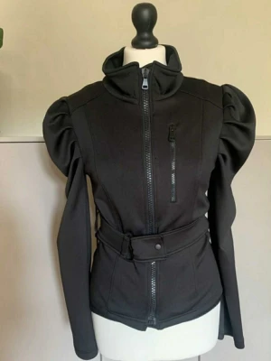 Svart jacka med puffärmar och bälte - Snygg svart jacka med puffiga axlar, hög krage och bälte i midjan. Jackan har dragkedja framtill och en bröstficka med dragkedja. Figurnära passform och långa ärmar ger en trendig siluett. Perfekt för dig som vill sticka ut.