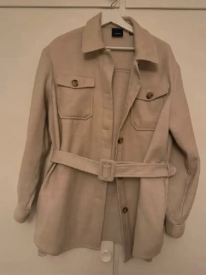Beige overshirt med bälte och fickor - Säljer en beige overshirt från only med bälte i midjan och två bröstfickor med knappar. Overshirten har klassisk krage och knäppning framtill. Perfekt att slänga över en t-shirt för en cool look.